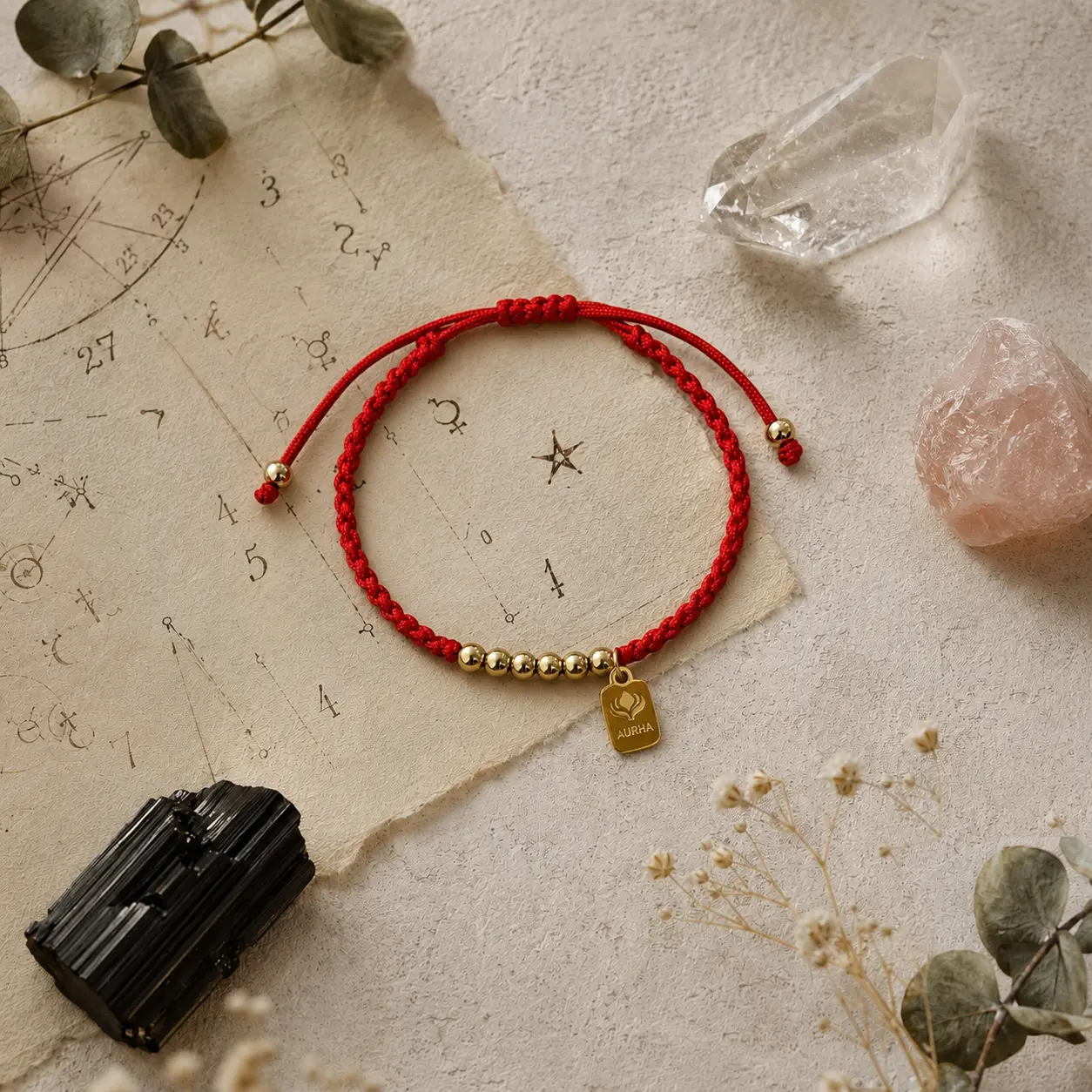 Pulseira Aurha vermelha com pingentes de estrela, bússola e lua, sobre carta astral com cristais de quartzo, quartzo rosa e turmalina negra